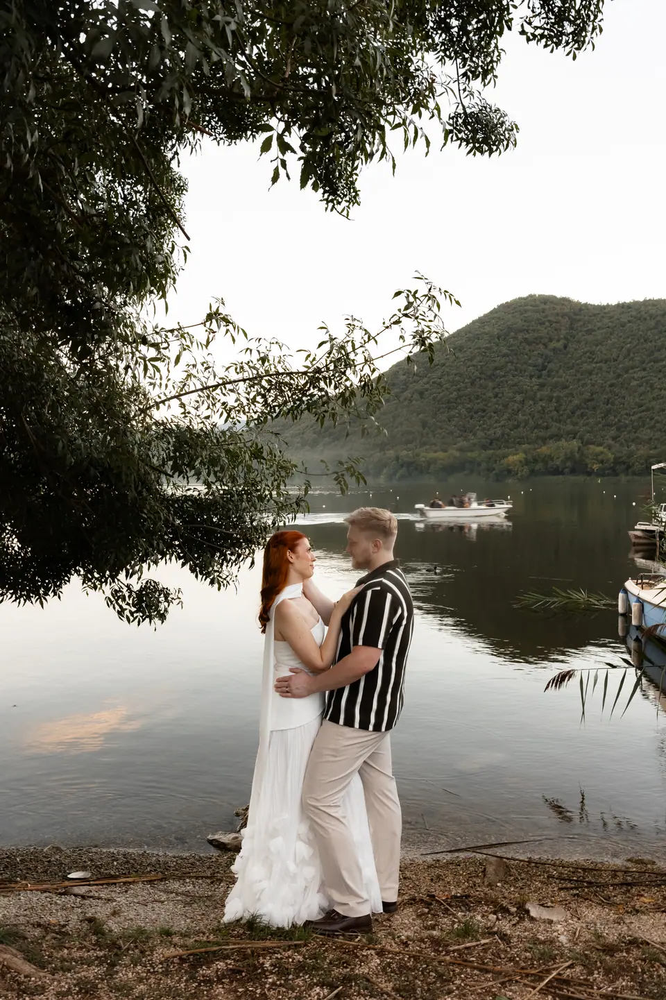 Kate and James embracing on the lake shore, wooded hills across the water