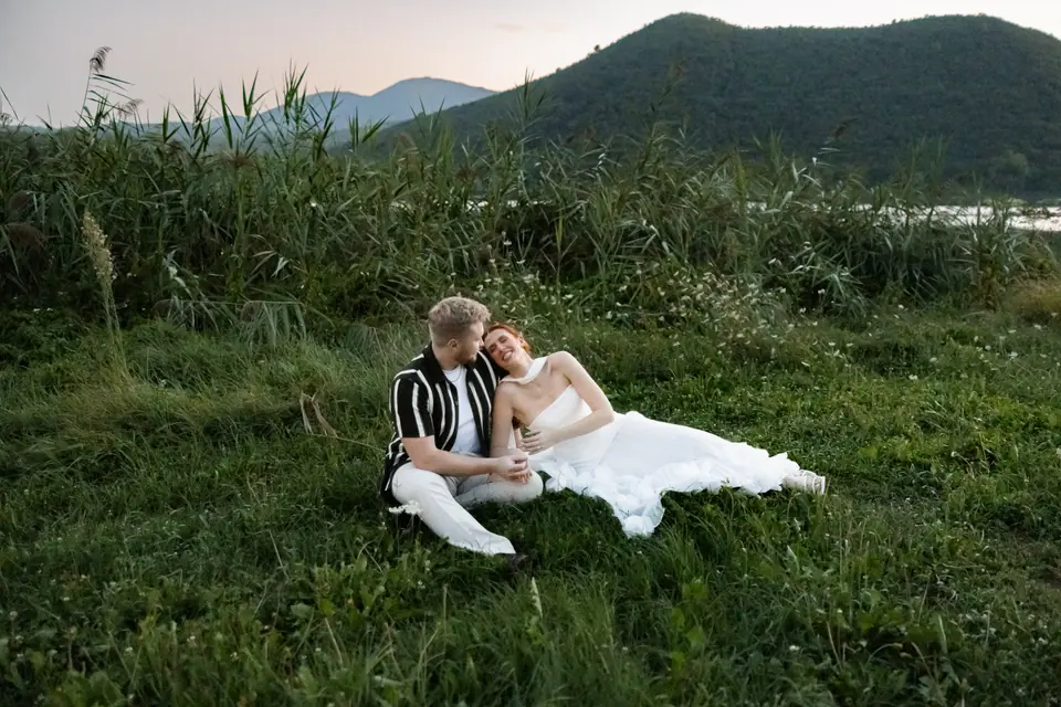 Kate and James lying in long grass by a lake with mountains behind