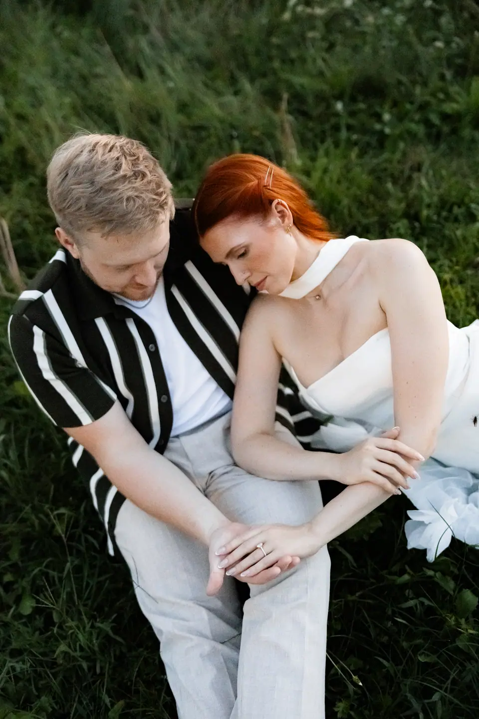 Kate resting against James in the grass, their hands and her ring together