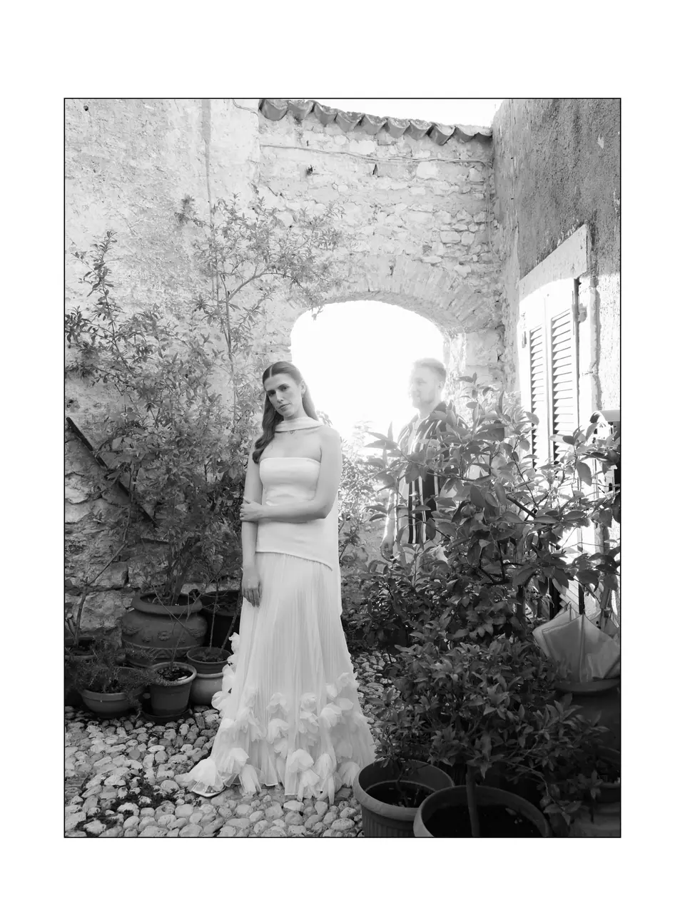 Black and white of Kate standing in a courtyard of plants, James behind her in the arch