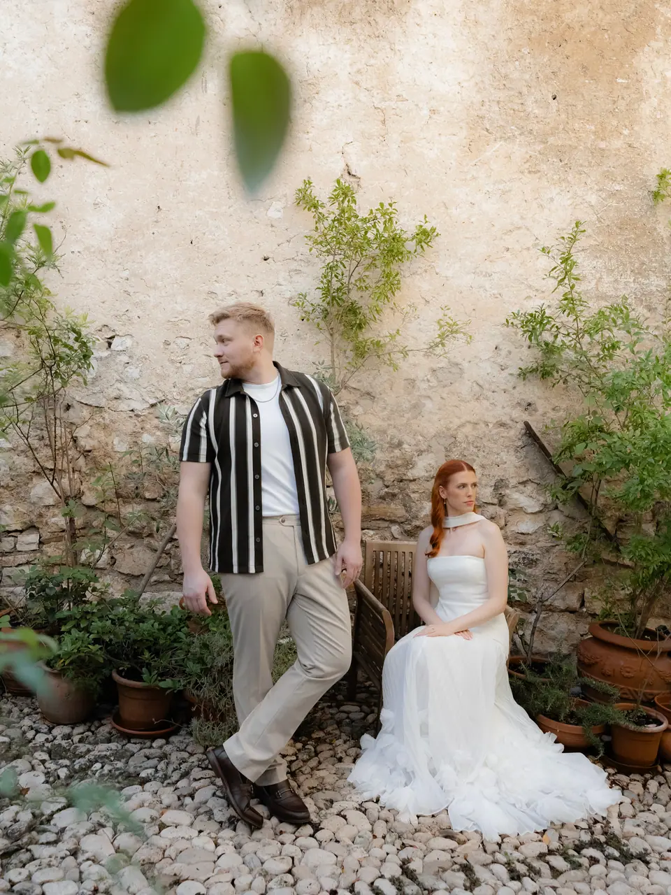 James standing beside Kate as she sits on a chair in a courtyard of potted plants