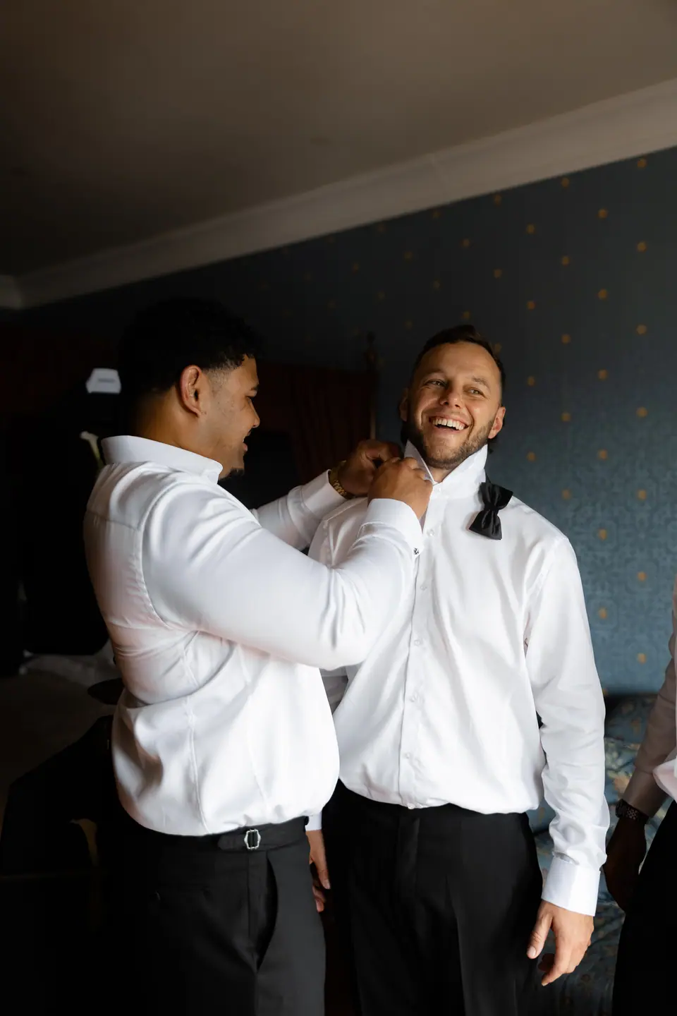 A groomsman fastening another’s bow tie as they laugh in a blue-papered room