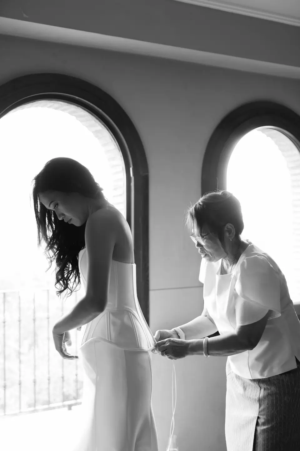 Black and white photograph of an older woman fastening Sivitha’s dress by the arched windows