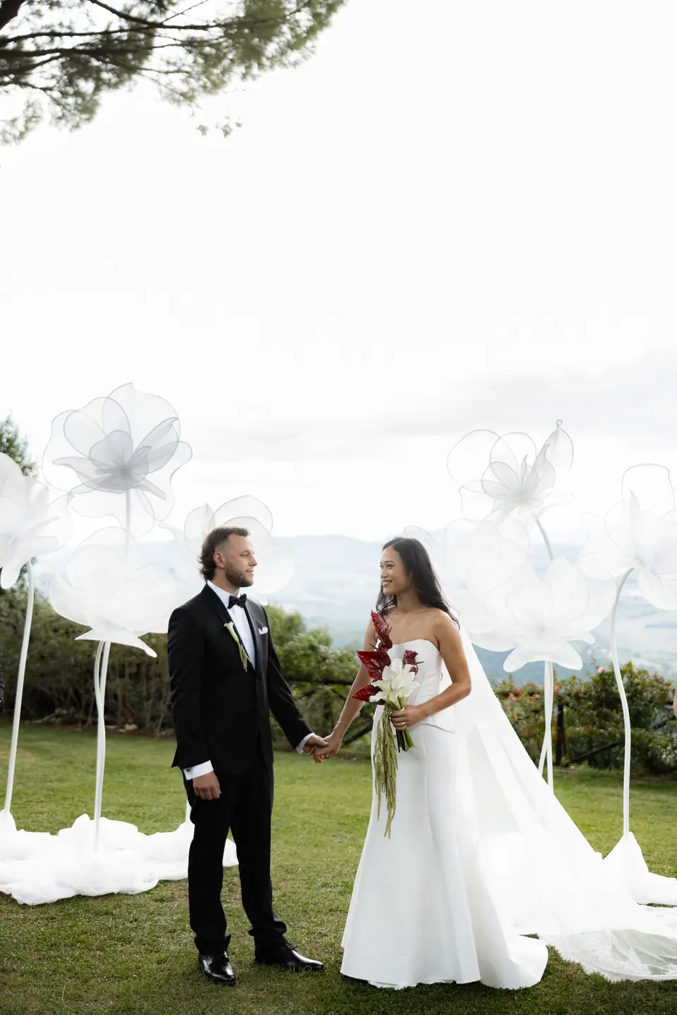 Sivitha and Rory holding hands among the paper flowers, her red bouquet in hand
