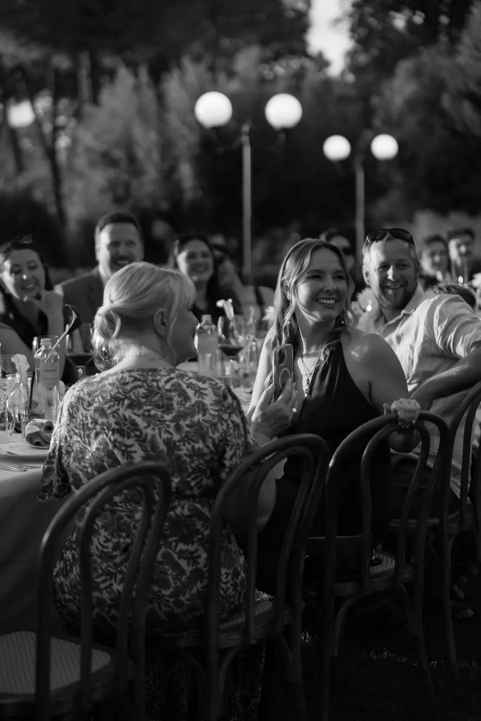 Black and white photograph of guests laughing at the long table under the lamps