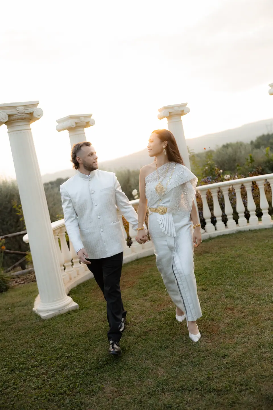 Sivitha in silver silk with a gold belt and Rory in white walking across the lawn
