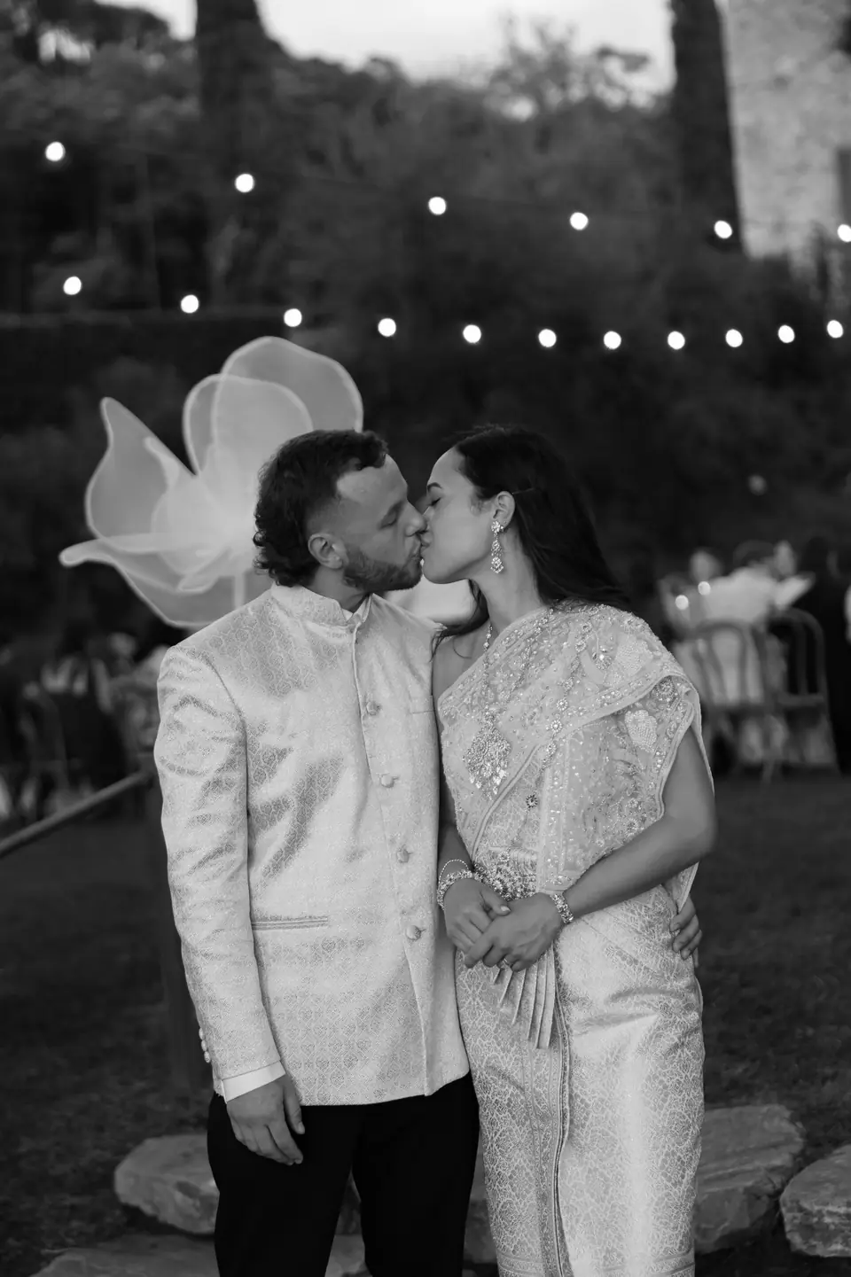 A black and white photograph of Sivitha and Rory kissing beneath string lights