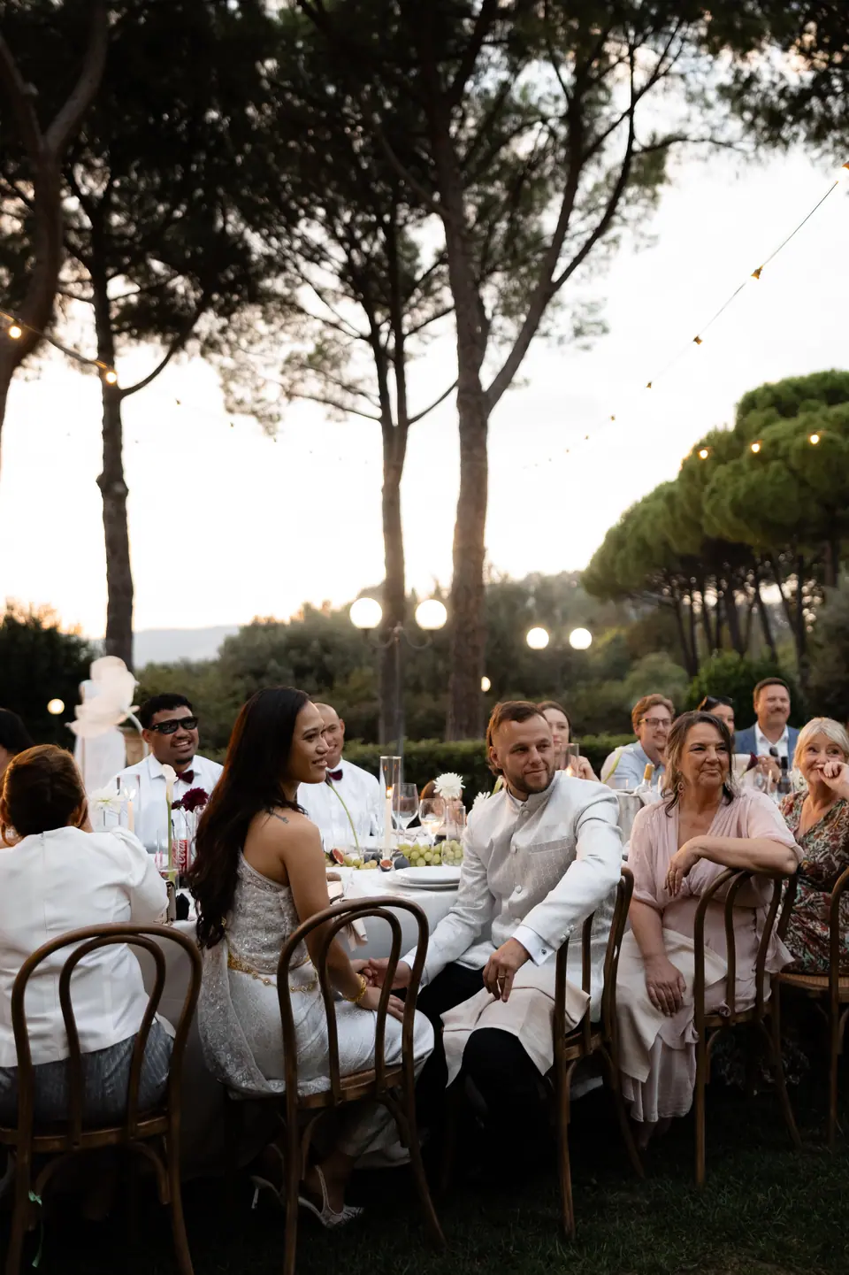 Guests at the long dinner table under the pines as dusk falls
