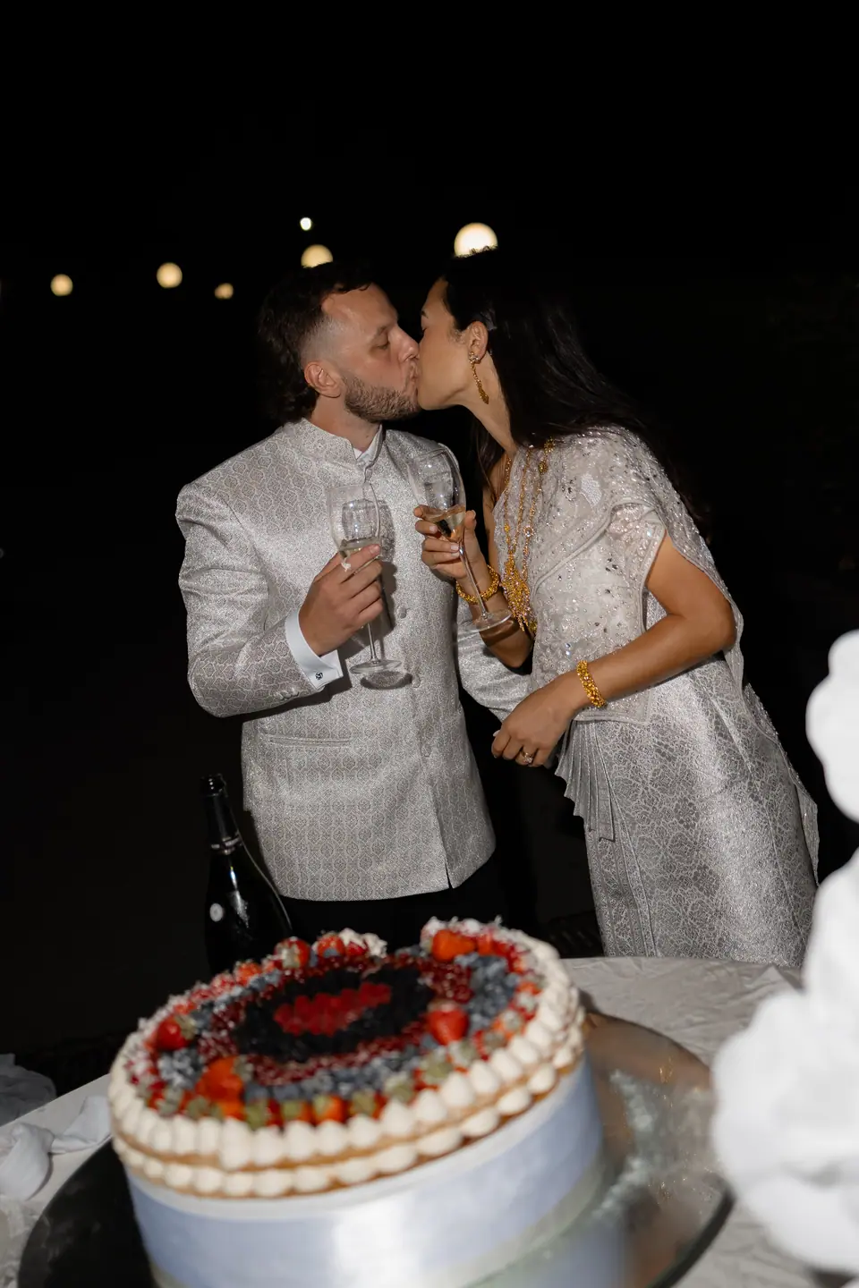 Sivitha and Rory kissing over glasses of champagne beside the cake