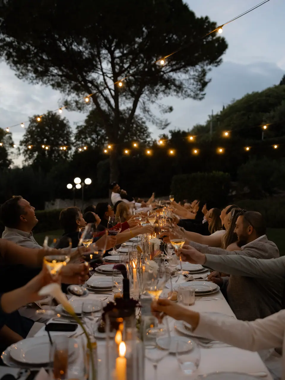 Guests raising glasses along the candlelit table beneath string lights and pines
