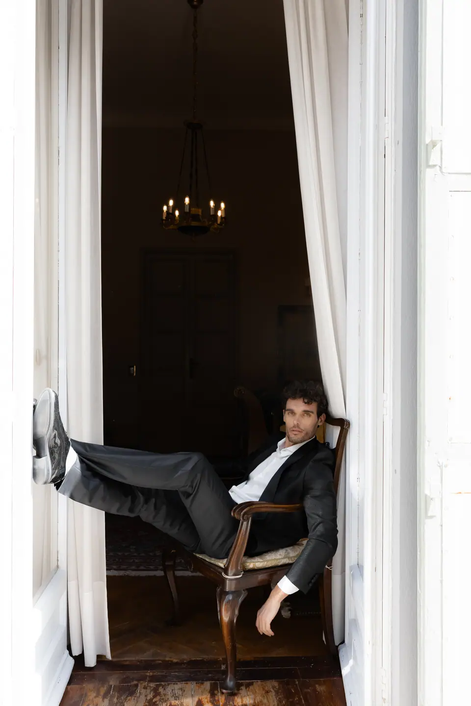Antoine seated in an armchair between white curtains, one foot raised against the doorframe
