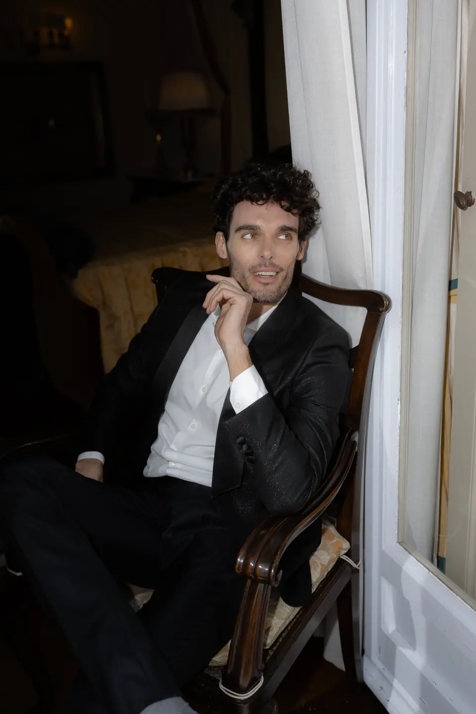 Antoine in a dinner jacket resting his chin on his hand in an armchair by the window