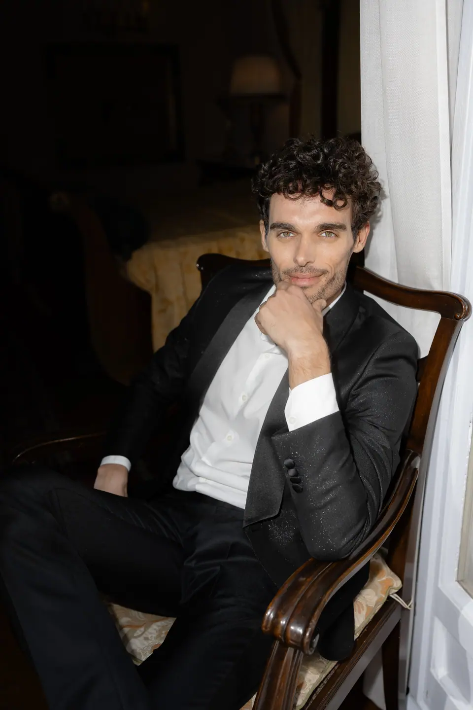 Antoine smiling at the camera, his chin on his hand, in an armchair by the window
