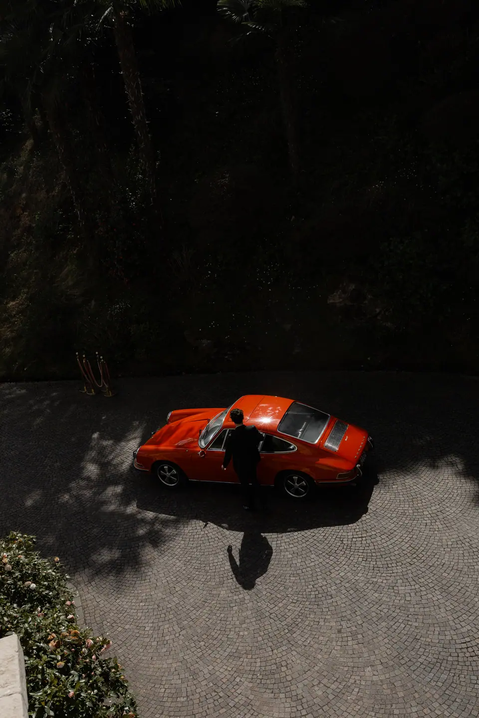 A red vintage Porsche seen from above, a figure standing beside it in the shade