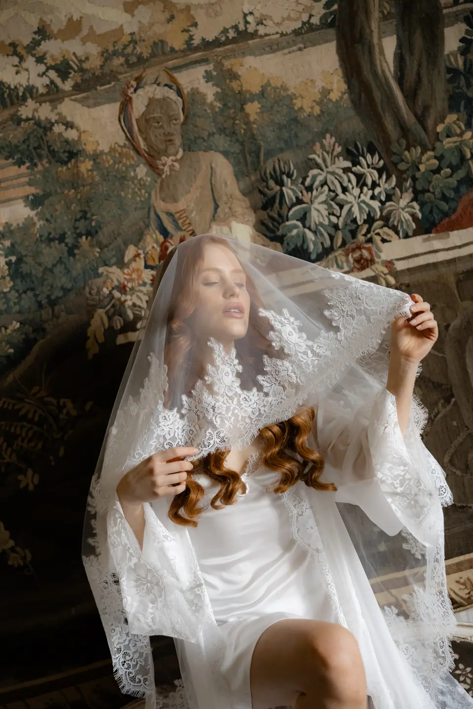 Roberta lifting a lace veil over her face in front of a tapestry
