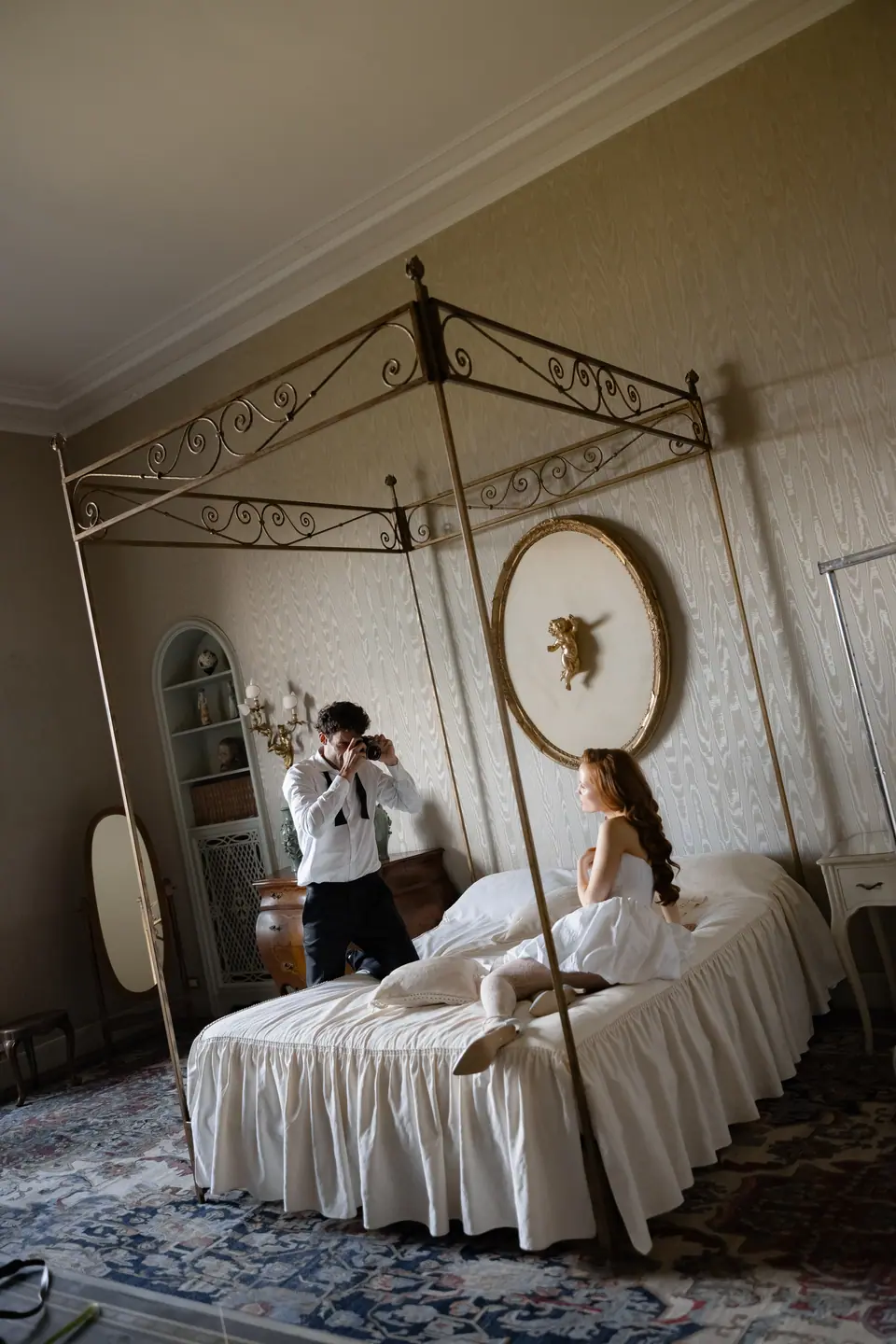 Antoine taking a photograph of Roberta sitting on a four-poster bed