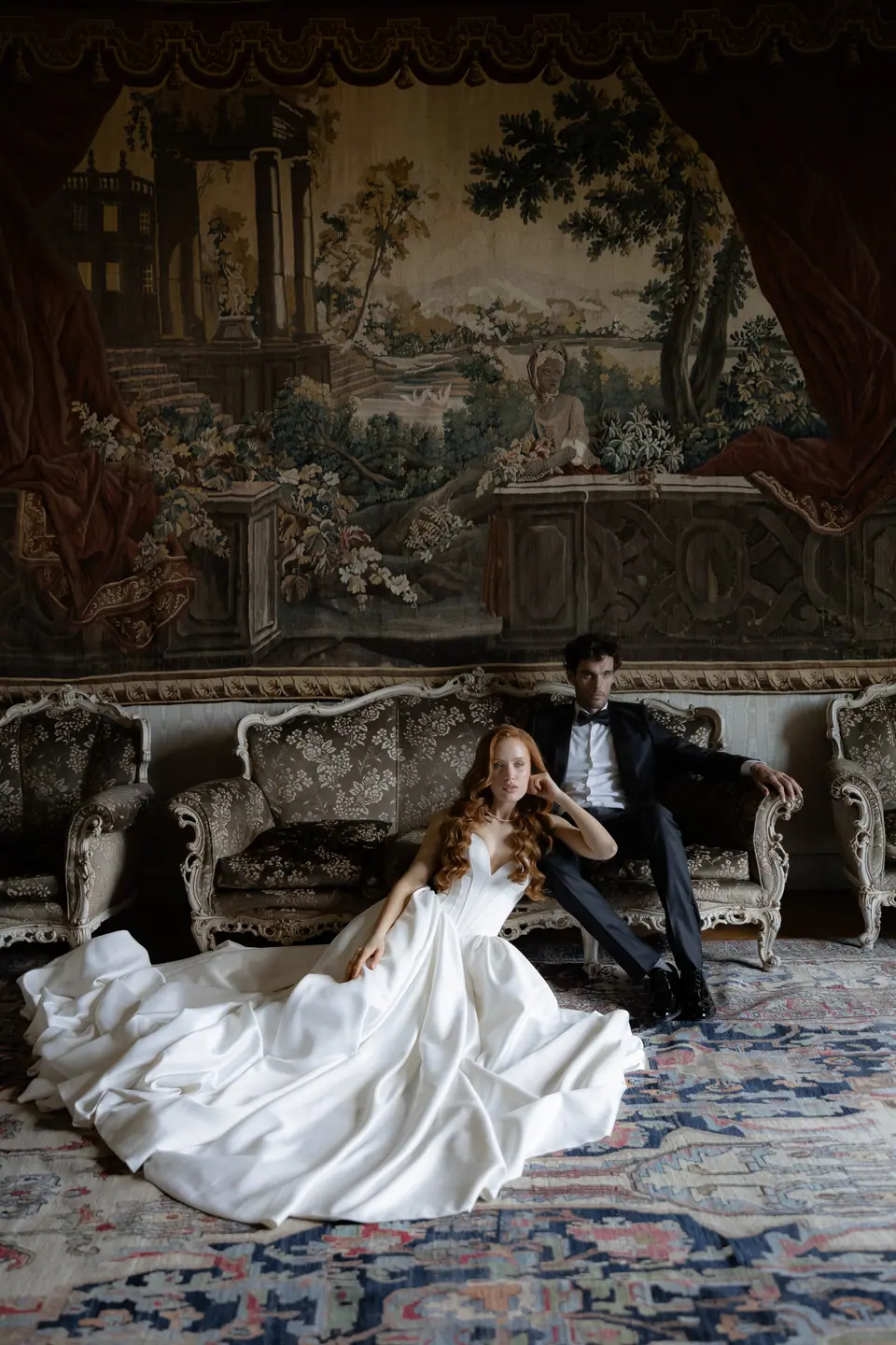 Roberta in a full white gown on a patterned rug before a tapestry, Antoine on the sofa behind her