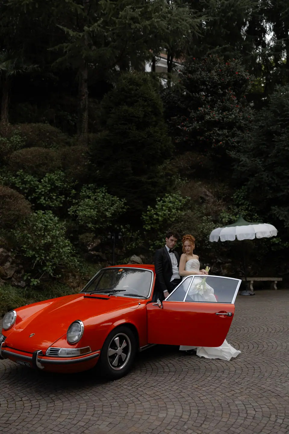 Roberta and Antoine beside a red vintage Porsche in the villa’s wooded garden