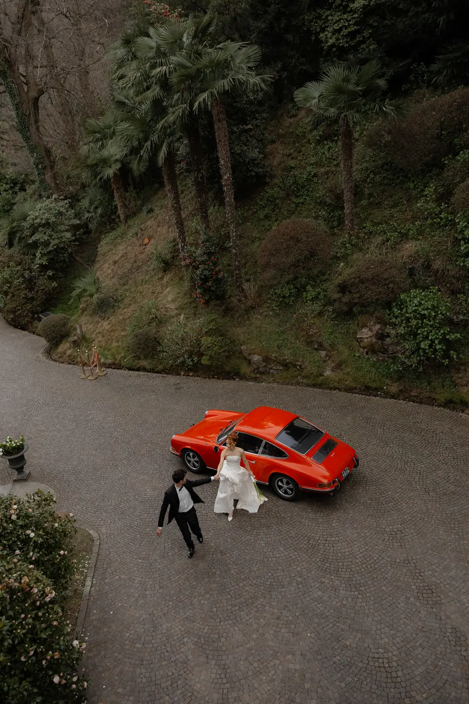 Roberta and Antoine beside the red Porsche on a winding drive, seen from above