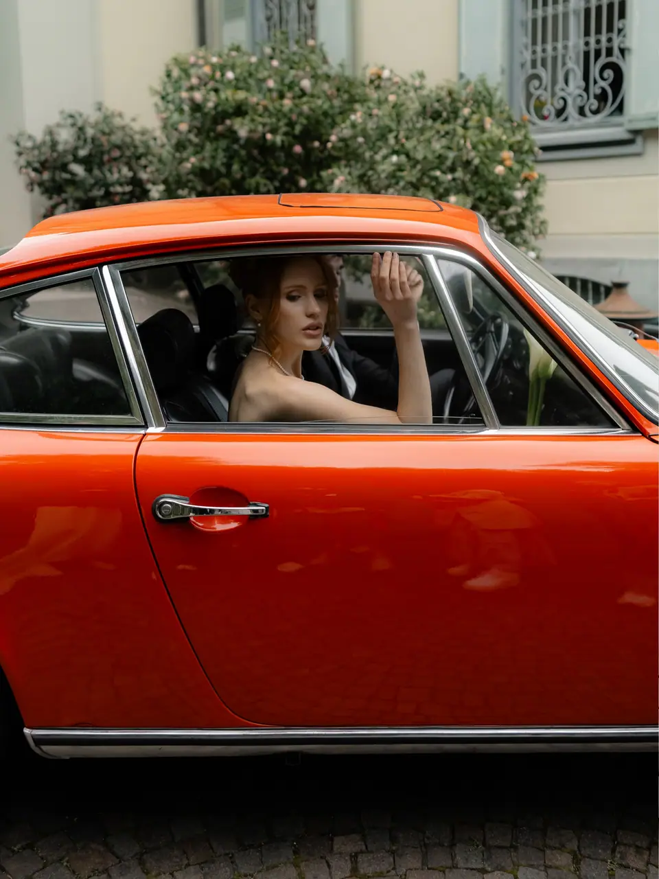 Roberta in the window of a red vintage Porsche, a rose bush behind