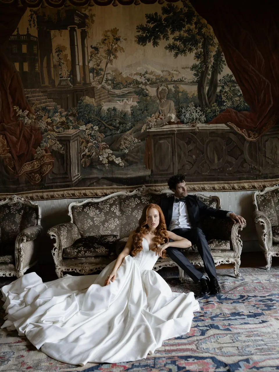 Roberta seated in her gown on a rug before a tapestry, Antoine on a sofa behind
